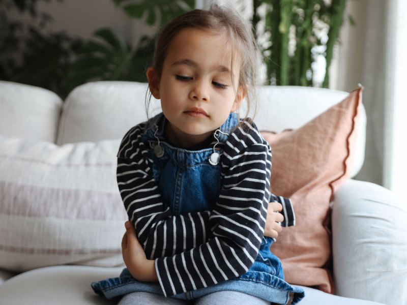 Mädchen, ca. 4 Jahre alt, sitzt mit verschränkten Armen auf dem Sofa und schaut traurig auf den Boden.