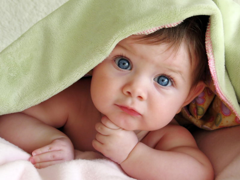 Baby mit blauen Augen liegt auf dem Boden und schaut unter der Decke hervor, direkt in die Kamera.
