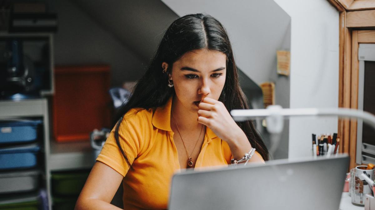 Junge Frau mit dunklen Haaren und senfgelben Shirt sitzt am Schreibtisch und schreibt eine Bewerbung an ihrem Laptop.