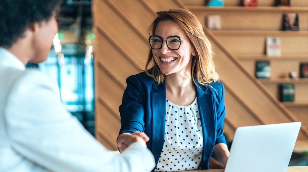 Frau sitzt auf der Arbeit an einem Tisch vor einem Laptop und gibt einer anderen Frau die Hand. Dabei lächelt sie stark.