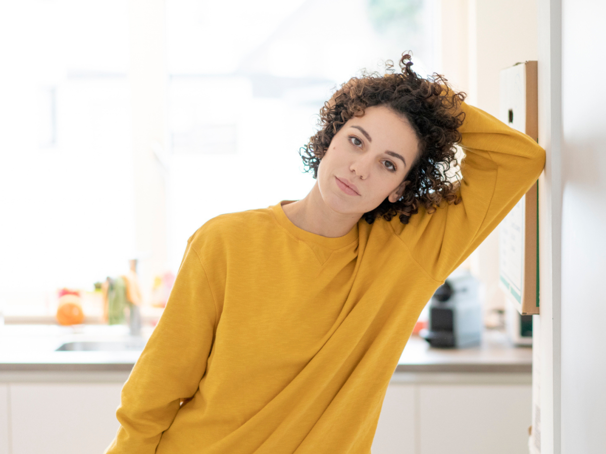 Frau mit dunklen Locken und einem gelben Pullover schaut ernst