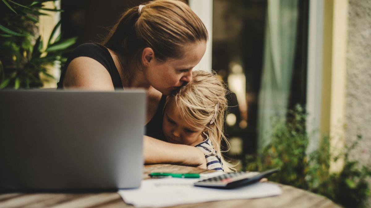 Frau arbeitet am Tisch und Küsst dabei ihre Tochter auf die Stirn, auf die sie neben der Arbeit aufpasst.