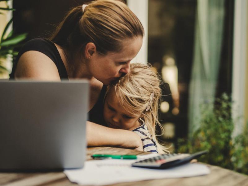 Frau arbeitet am Tisch und Küsst dabei ihre Tochter auf die Stirn, auf die sie neben der Arbeit aufpasst.
