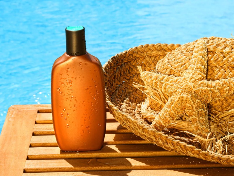 Eine Tube Sonnencreme vor einem blauen Pool und neben einem Sonnenhut.