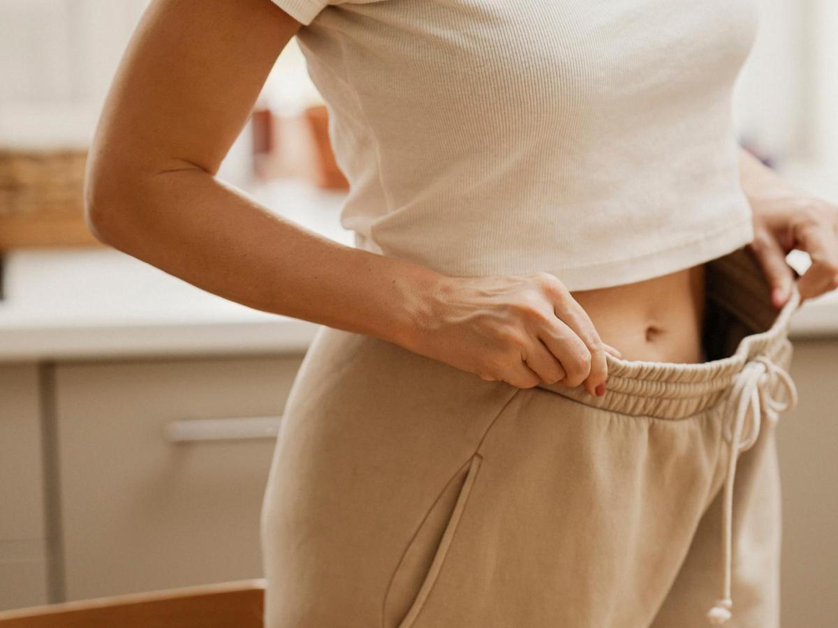 Frau hat eine zu weite Jogginghose mit Gummizug an und zeigt mit den Händen, wie viel Platz die Hose oben am Bund hat.