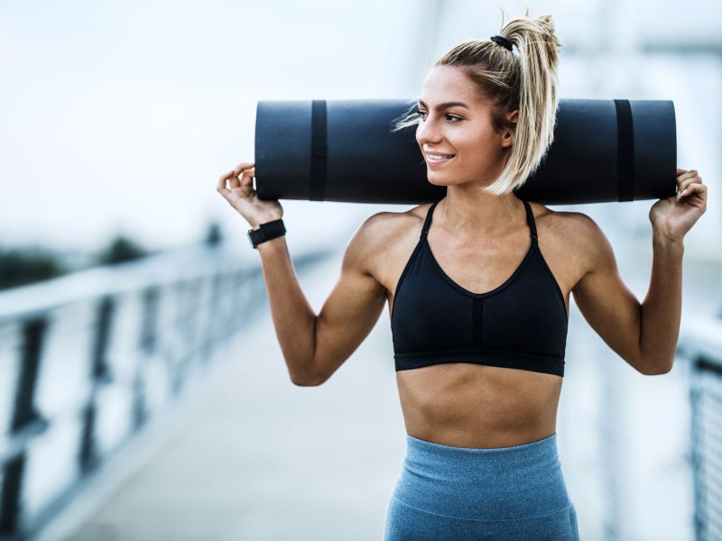 Sportliche Frau mit trainiertem Körper trägt Sportmatte über den Schultern