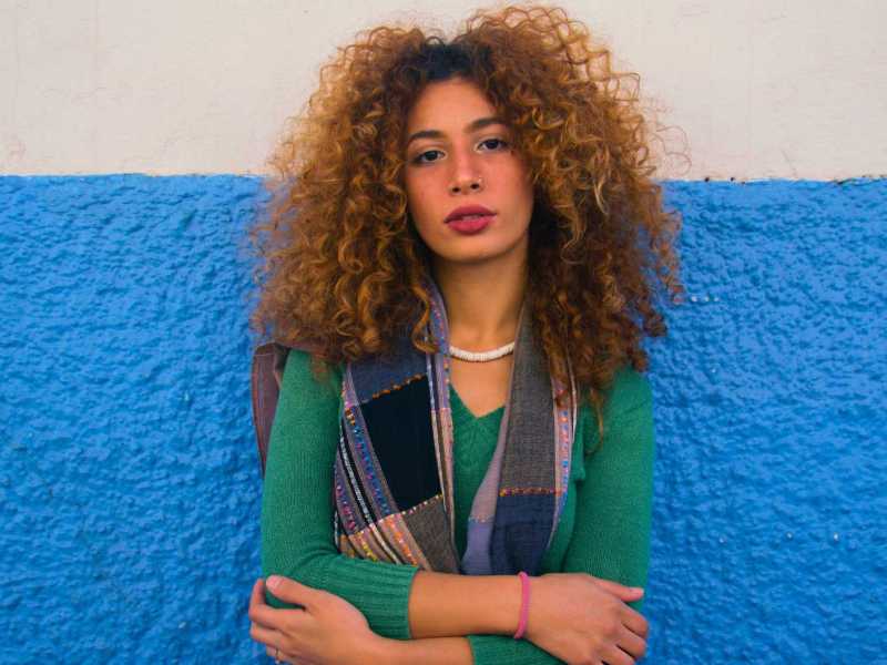 Junge Frau mit Locken steht vor einer blau-weißen Wand und schaut in die Kamera.