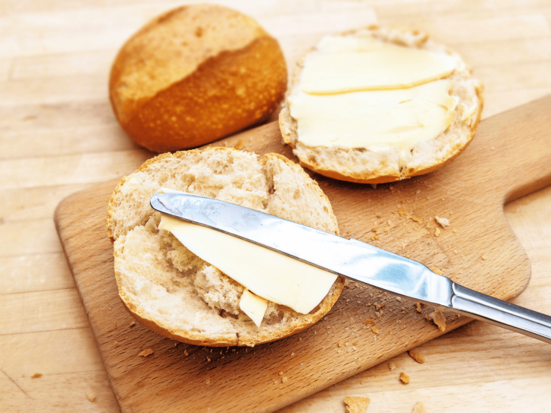 Aufgeschnittenes Brötchen auf einem Holzbrettchen, mit Butter bestrichen.