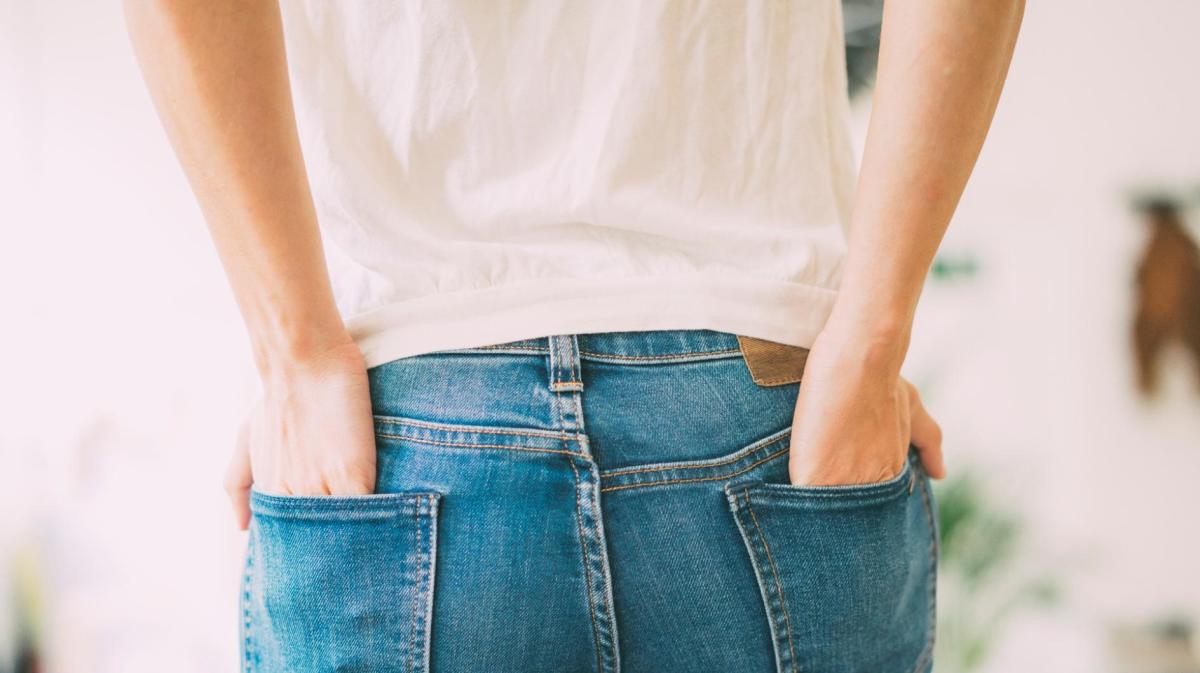 Frau steht mit dem Rücken zur Kamera und hat beide Hände in den hinteren Hosentaschen ihrer Jeans.
