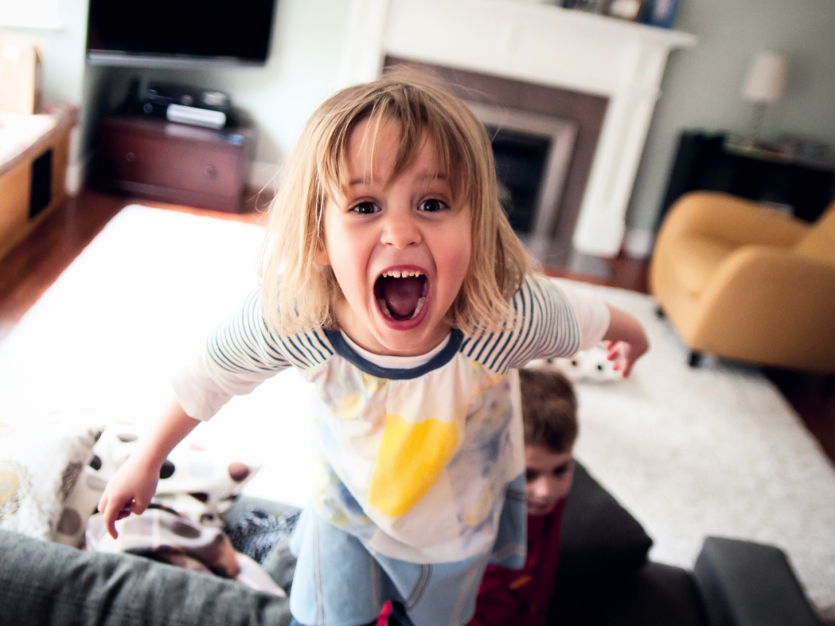 Kind mit blonden Haaren steht im Wohnzimmer auf der Couch und schreit in die Kamera.