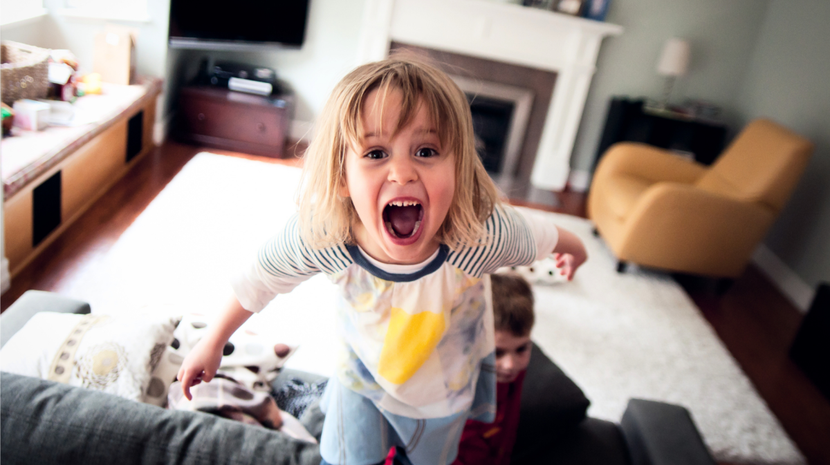 Kind mit blonden Haaren steht im Wohnzimmer auf der Couch und schreit in die Kamera.