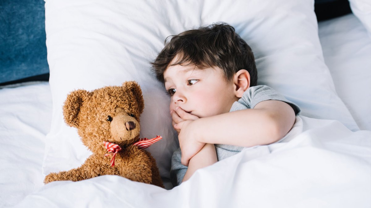 Kleines Kind liegt im Bett und schaut seinen Plüschteddy an, der neben ihm liegt.