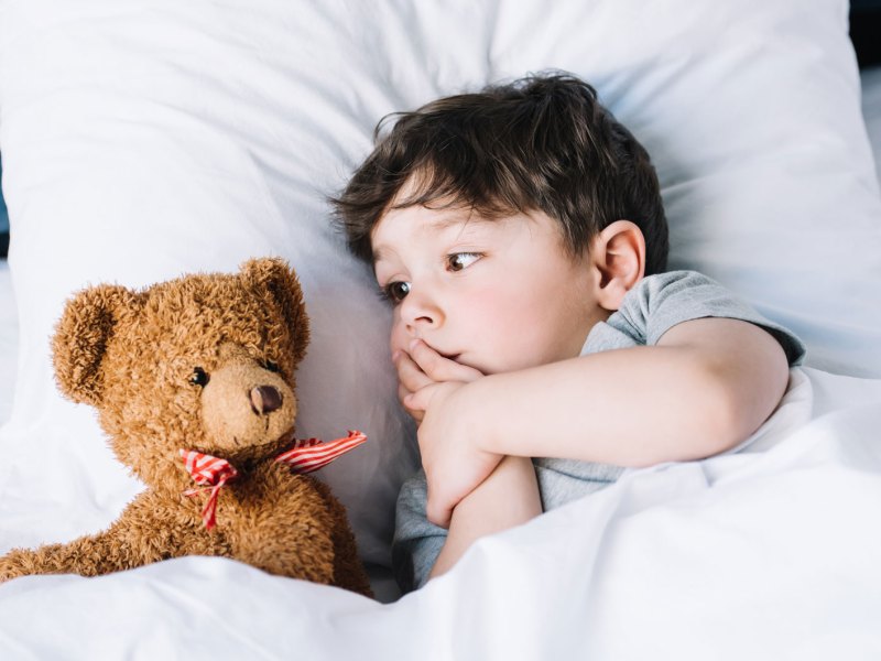 Kleines Kind liegt im Bett und schaut seinen Plüschteddy an, der neben ihm liegt.