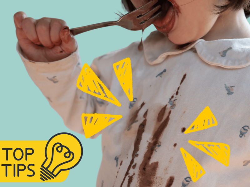 Kind isst mit einer Gabel Schokoladensoße und kleckert auf die Bluse, Fokus mit gelben Zeichen auf die Flecken.