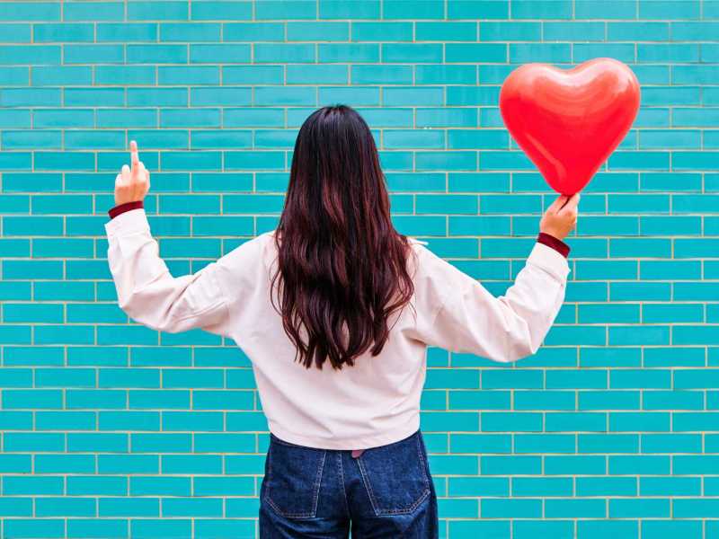 Junge Frau zeigt Mittelfinger und hält roten Ballon vor türkiser Ziegelwand.