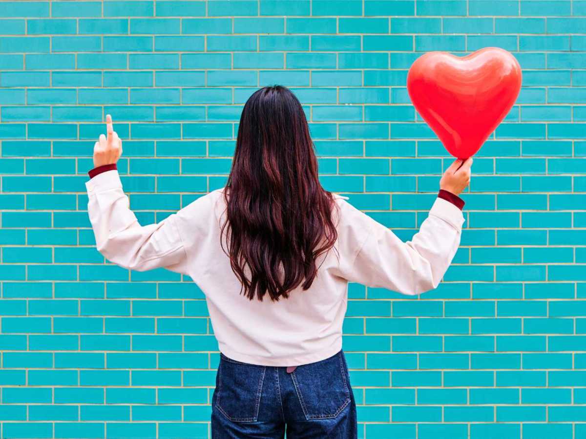 Junge Frau zeigt Mittelfinger und hält roten Ballon vor türkiser Ziegelwand.