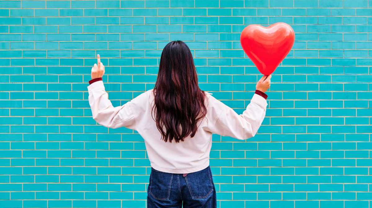 Junge Frau zeigt Mittelfinger und hält roten Ballon vor türkiser Ziegelwand.