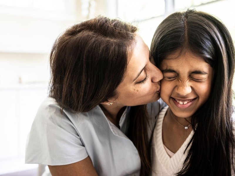 Mutter gibt ihrer Tochter einen Kuss auf die Wange, die verzieht witzig das Gesicht.