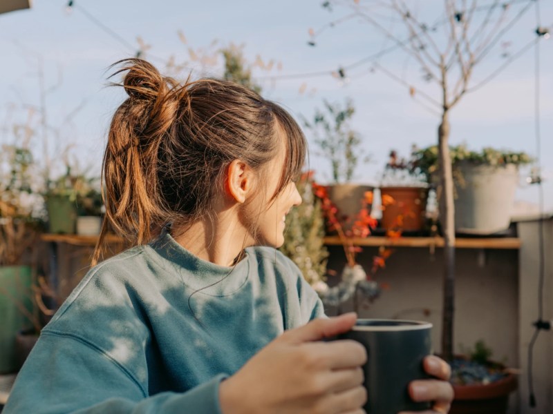 junge Frau sitzt auf ihrem Balkon mit einer Kaffeetasse in der Hand