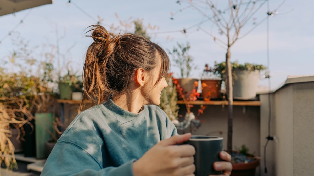 junge Frau sitzt auf ihrem Balkon mit einer Kaffeetasse in der Hand