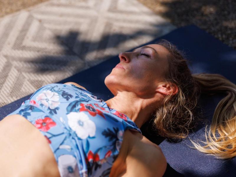 Frau mit blonden Haaren liegt auf einer Yogamatte im Schatten.