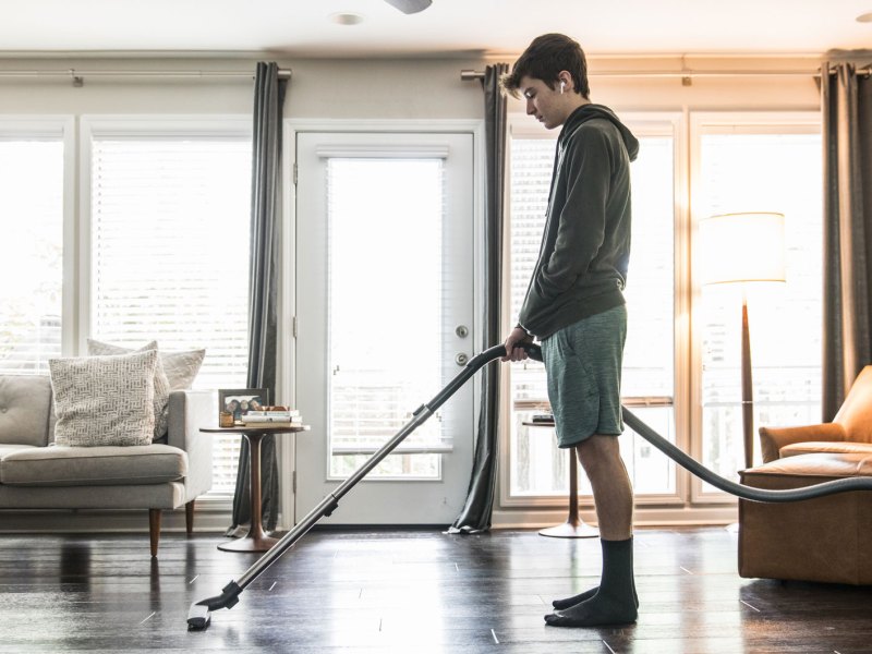 Männlicher Teenager steht unmotiviert mit Staubsauger im Wohnzimmer.