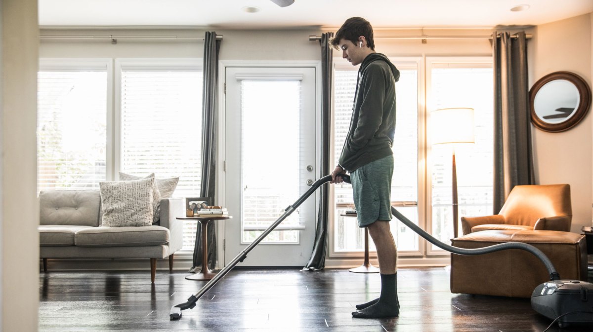 Männlicher Teenager steht unmotiviert mit Staubsauger im Wohnzimmer.