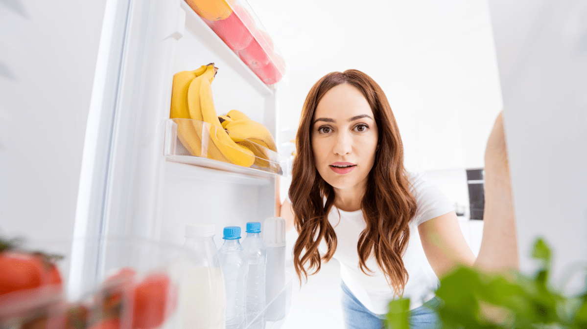 Eine dunkelhaarige Frau guckt in einen offenen Kühlschrank.