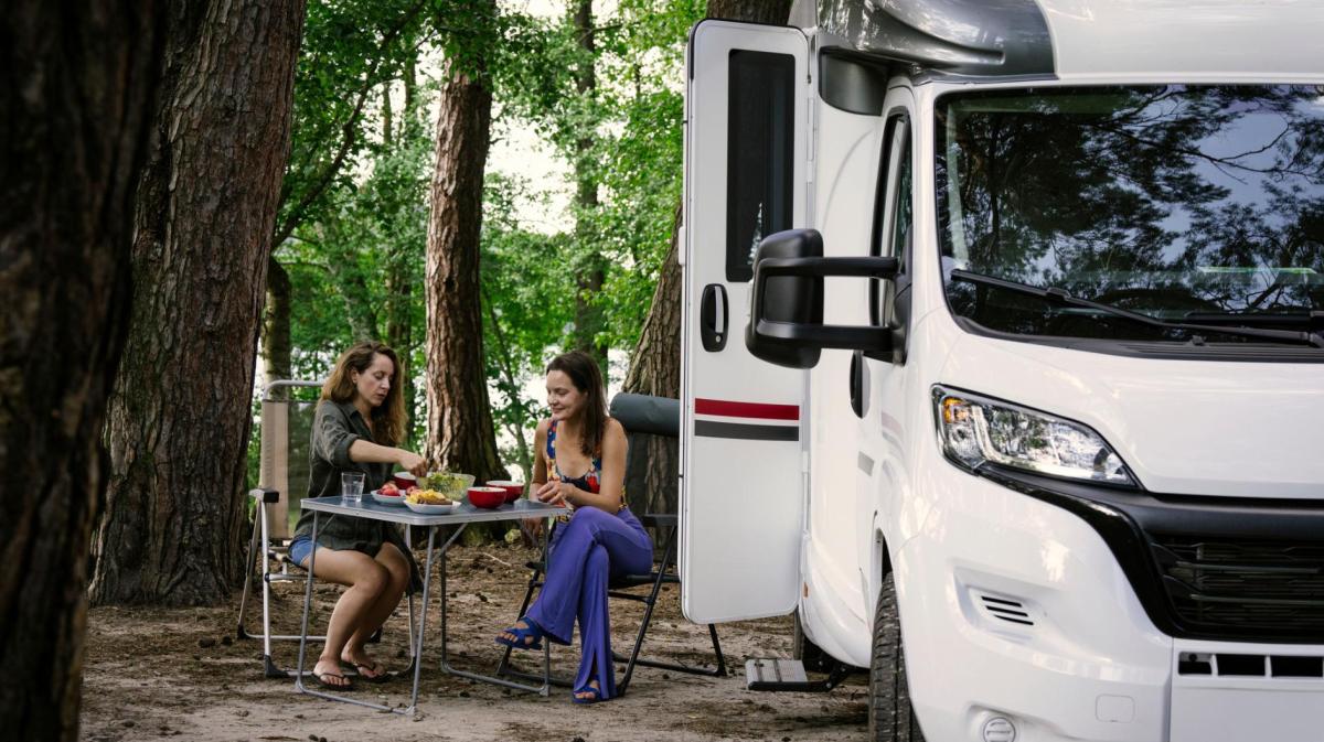 Zwei Frauen sitzen an einem Campingtisch in der Natur neben einem Wohnmobil.