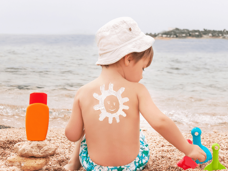 Kleiner Junge sitzt in Badekleidung am Strand und hat auf dem Rücken eine Sonne aus Sonnencreme gemalt