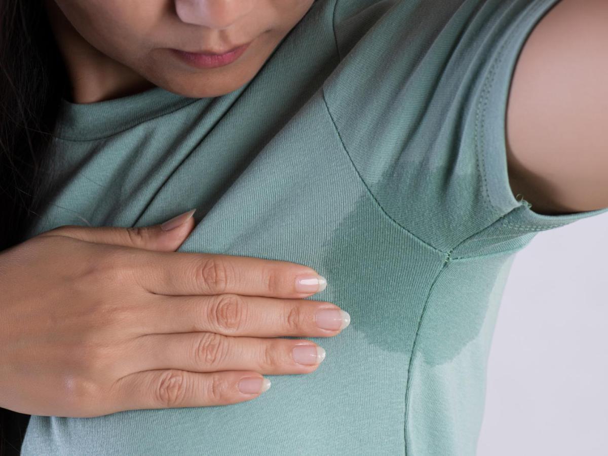Frau guckt unter ihre Achsel. Sie hat einen Schweißfleck, der auf dem grünen Shirt deutlich sichtbar ist.