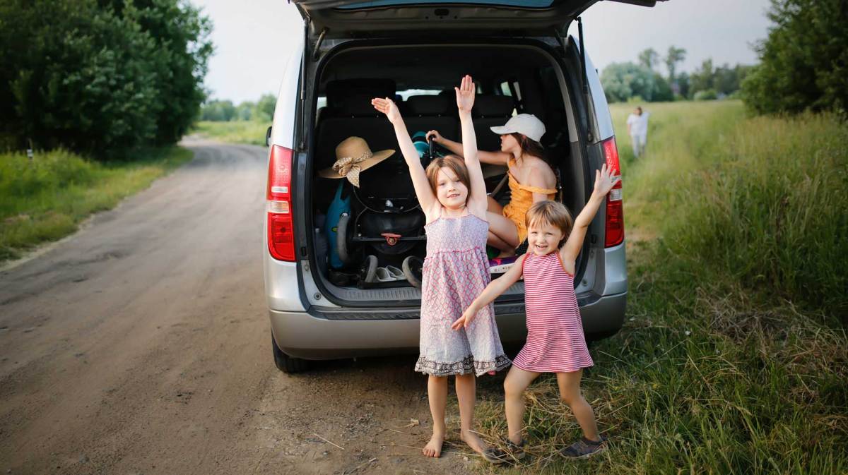 Zwei Kinder stehen vor dem vollen Kofferraum ihres Minivans. Ein weiteres Kind sitzt zwischen den Koffern.