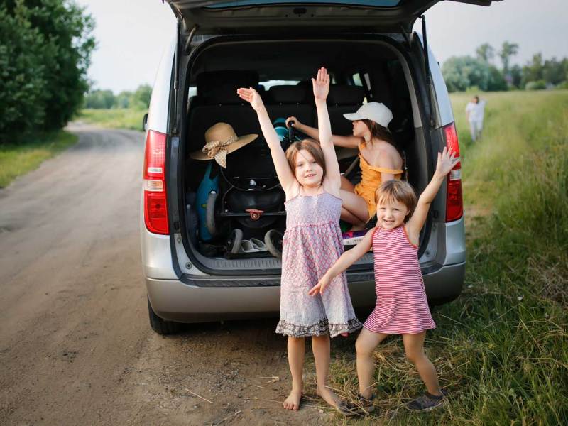 Zwei Kinder stehen vor dem vollen Kofferraum ihres Minivans. Ein weiteres Kind sitzt zwischen den Koffern.