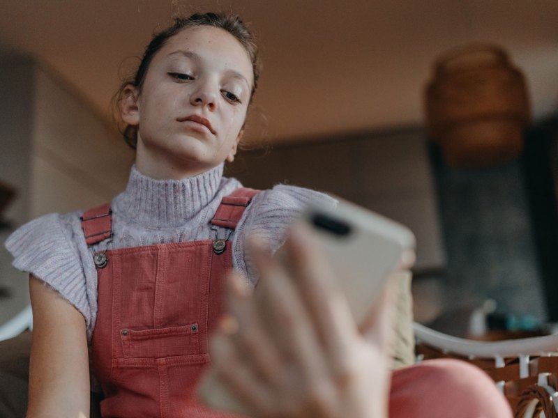 M&auml;dchen im Teenageralter sitzt auf dem Sofa und schaut genervt auf ihr Smartphone.