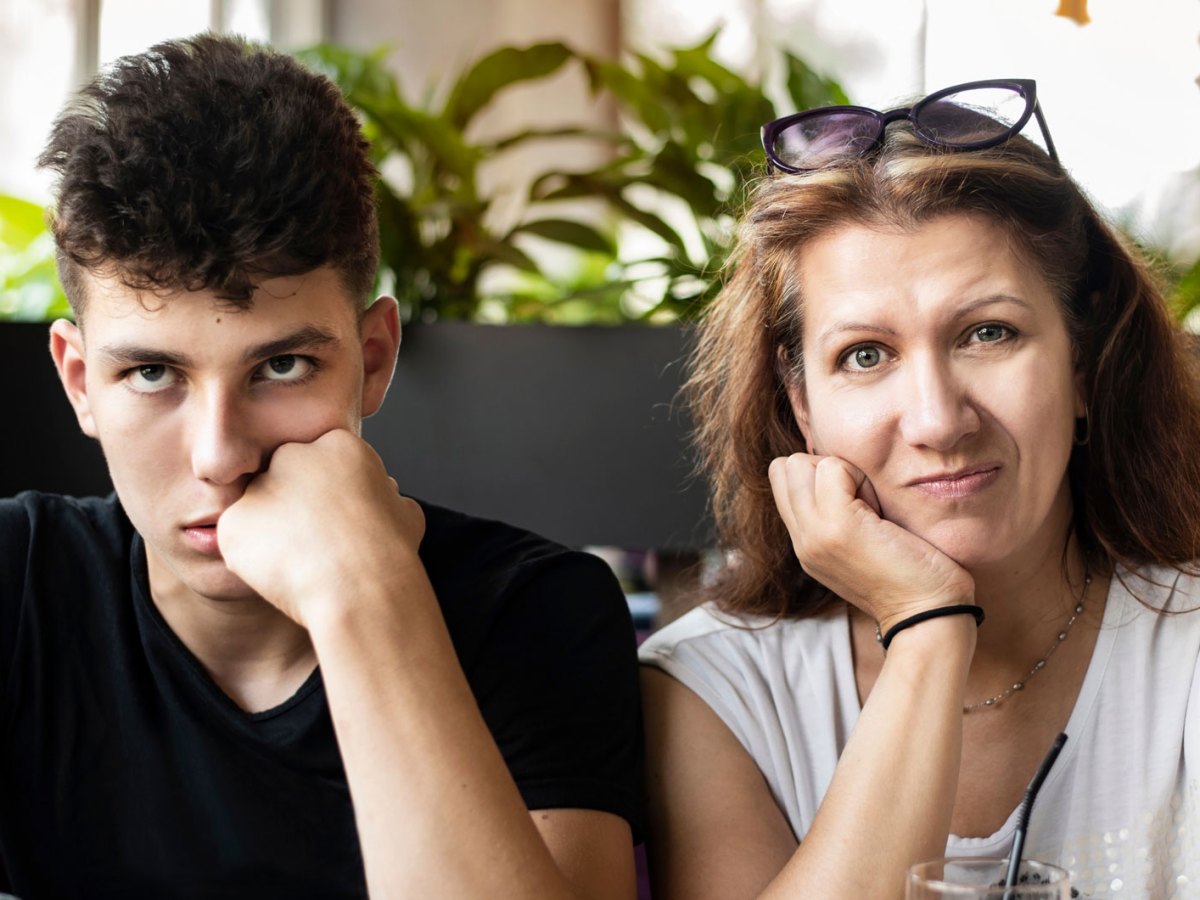 Frau mit ihrem Sohn im Teenageralter in einem Cafe. Der Sohn verdreht die Augen, die Mutter schaut verzweifelt.