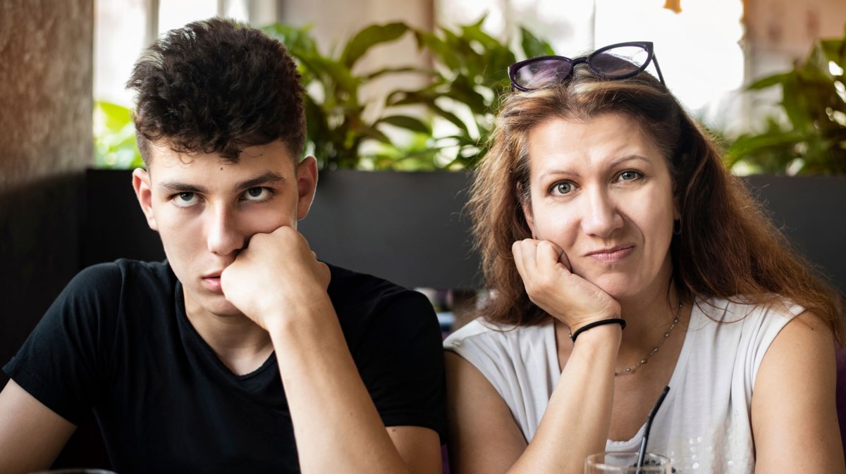 Frau mit ihrem Sohn im Teenageralter in einem Cafe. Der Sohn verdreht die Augen, die Mutter schaut verzweifelt.