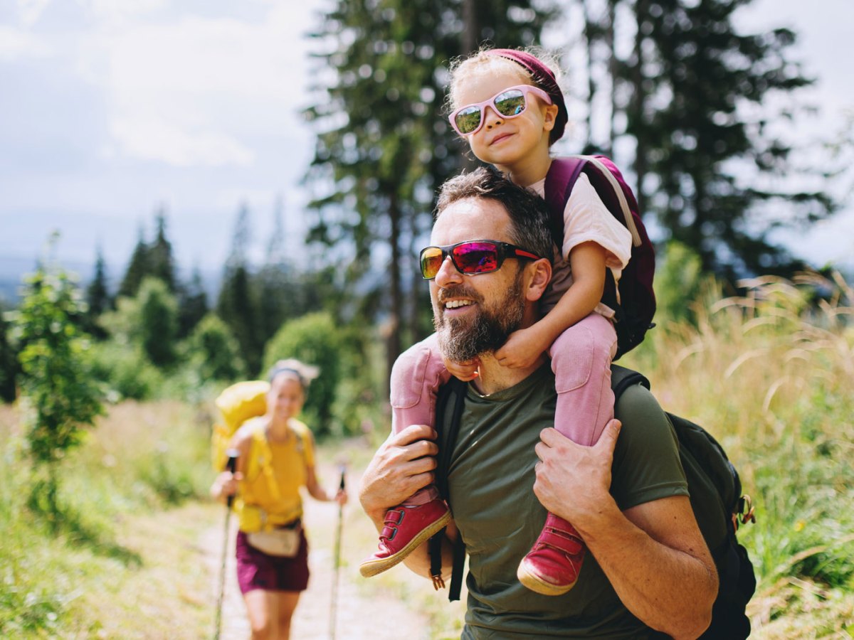 Eltern gehen mit ihrem Kind wandern. Der Vater tr&auml;gt die Tochter auf den Schultern.