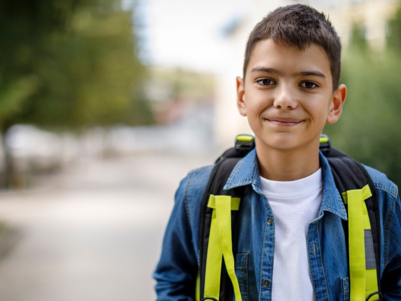 Porträt eines Jungen, ca. 10 Jahre alt, steht mit einem Rucksack auf der Straße und schaut freundlich in die Kamera.