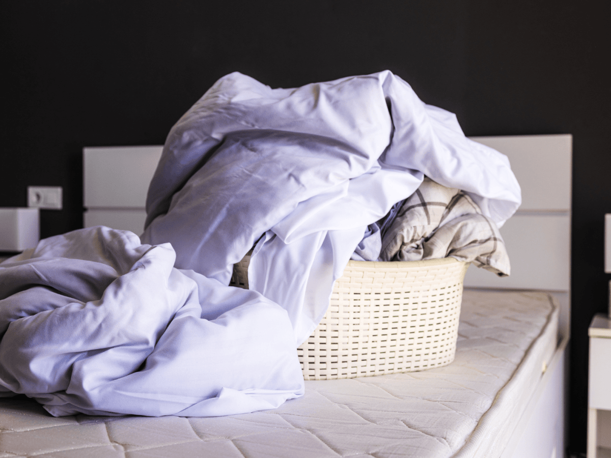 W&auml;schekorb mit Bettw&auml;sche steht auf einem abgezogenen Bett.