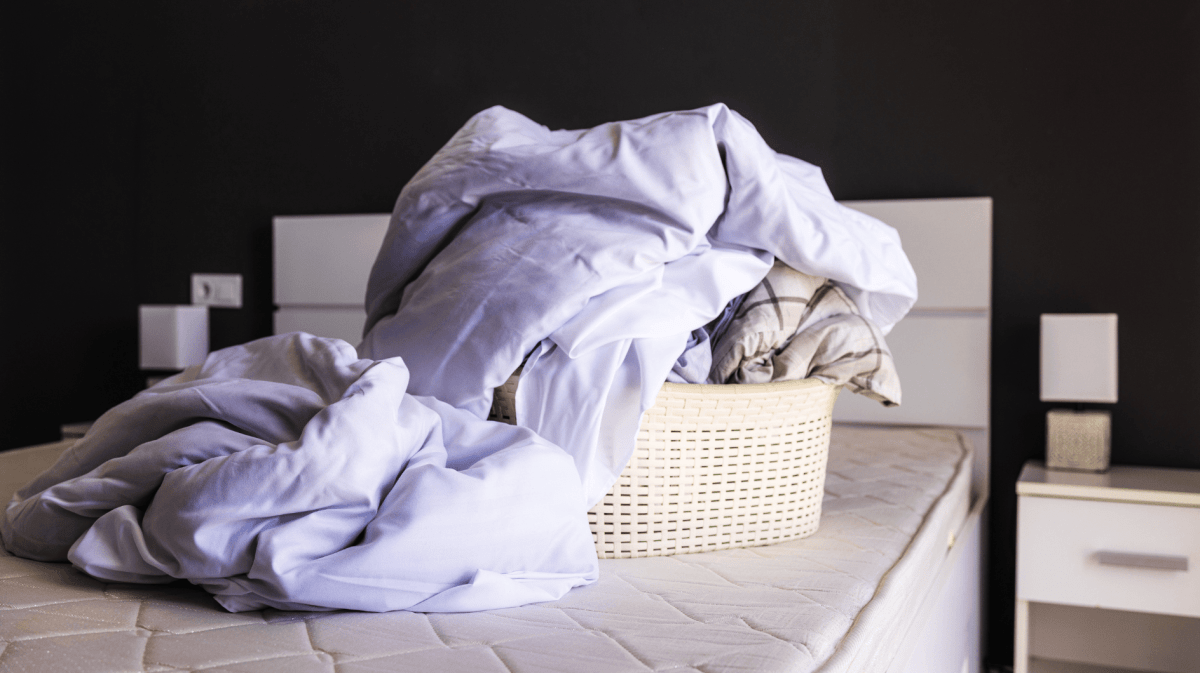 Wäschekorb mit Bettwäsche steht auf einem abgezogenen Bett.