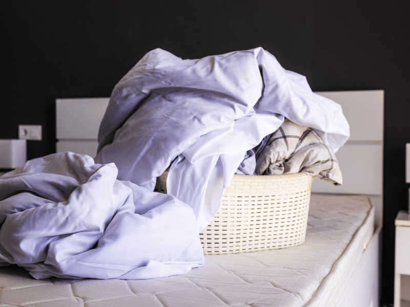 Wäschekorb mit Bettwäsche steht auf einem abgezogenen Bett.