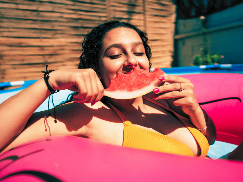 Frau sitzt mit einem pinken Schwimmreifen im Pool und isst eine Wassermelone.