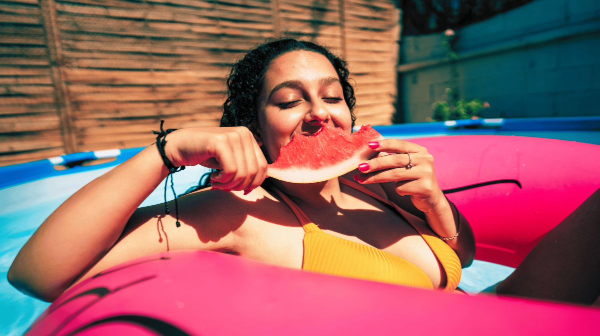 Frau sitzt mit einem pinken Schwimmreifen im Pool und isst eine Wassermelone.