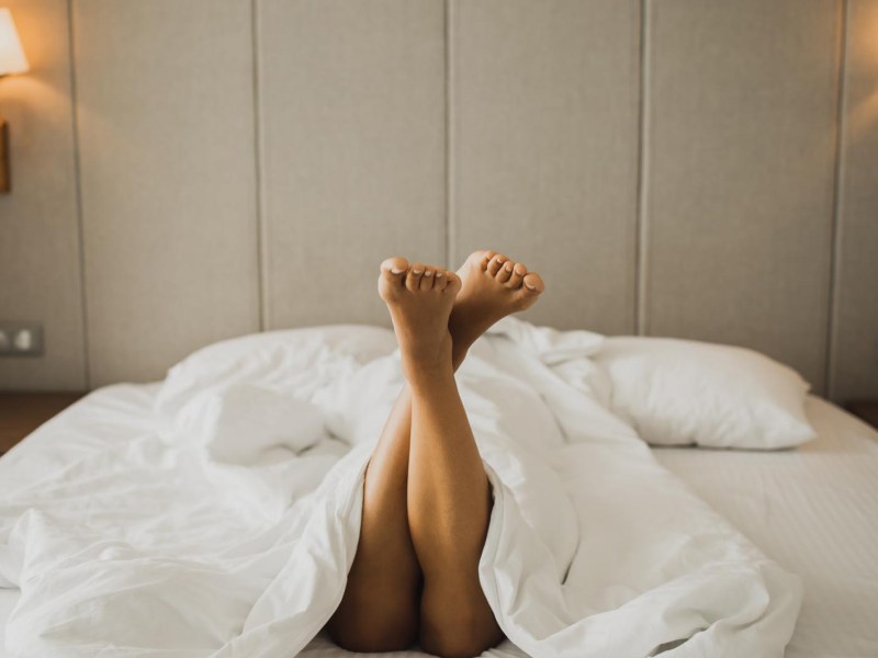 Frau liegt in einem Hotelbett, nackte Beine gucken unter der Decke hervor