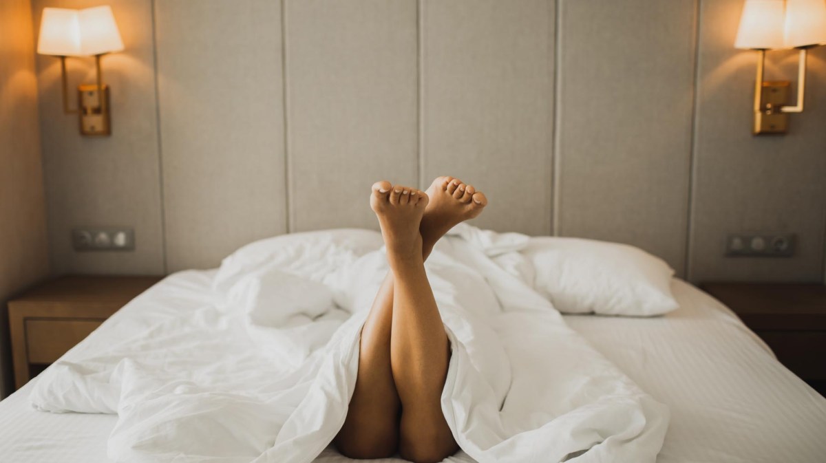 Frau liegt in einem Hotelbett, nackte Beine gucken unter der Decke hervor