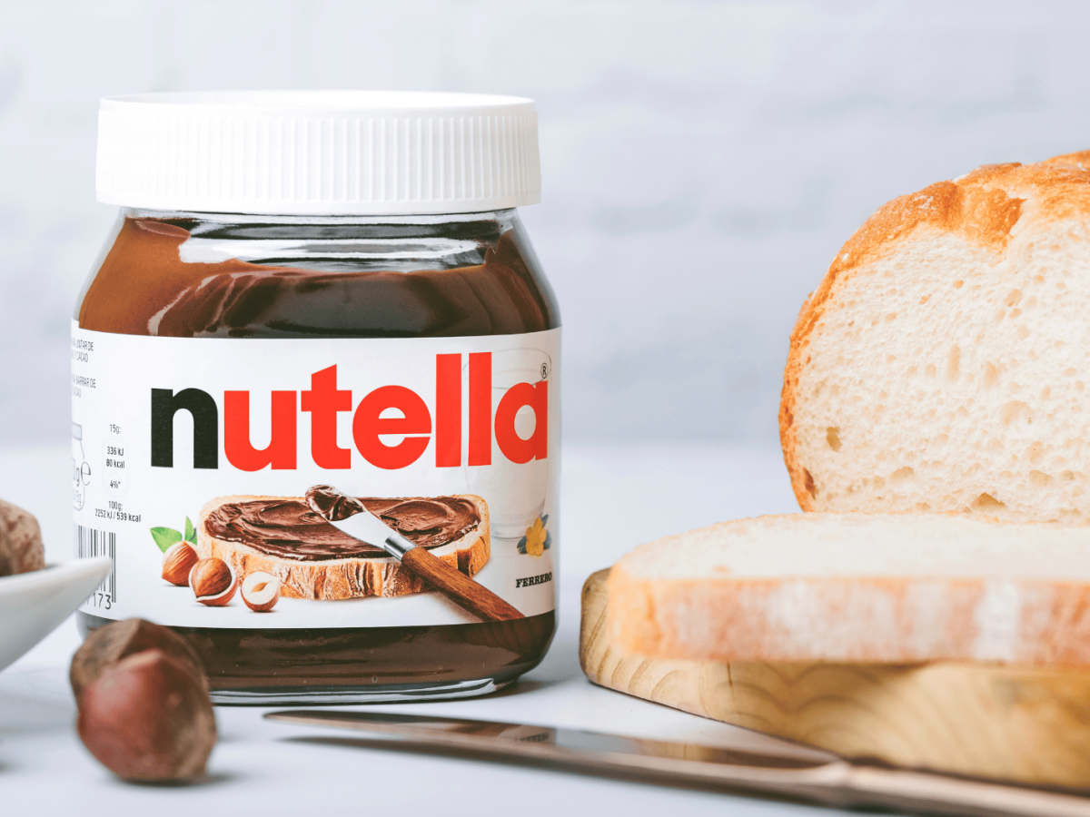 Nutella-Glas auf Tisch, neben Wei&szlig;brot und Haseln&uuml;ssen.