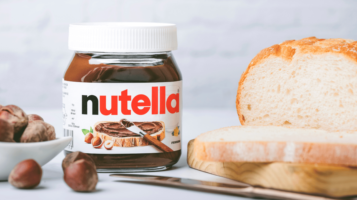 Nutella-Glas auf Tisch, neben Weißbrot und Haselnüssen.