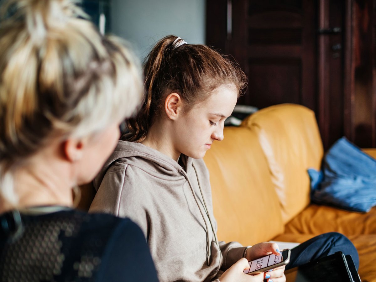 Mutter und Tochter auf der Couch. Die Tochter ist in ihr Handy vertieft, die Mutter versucht mimt ihr zu sprechen.