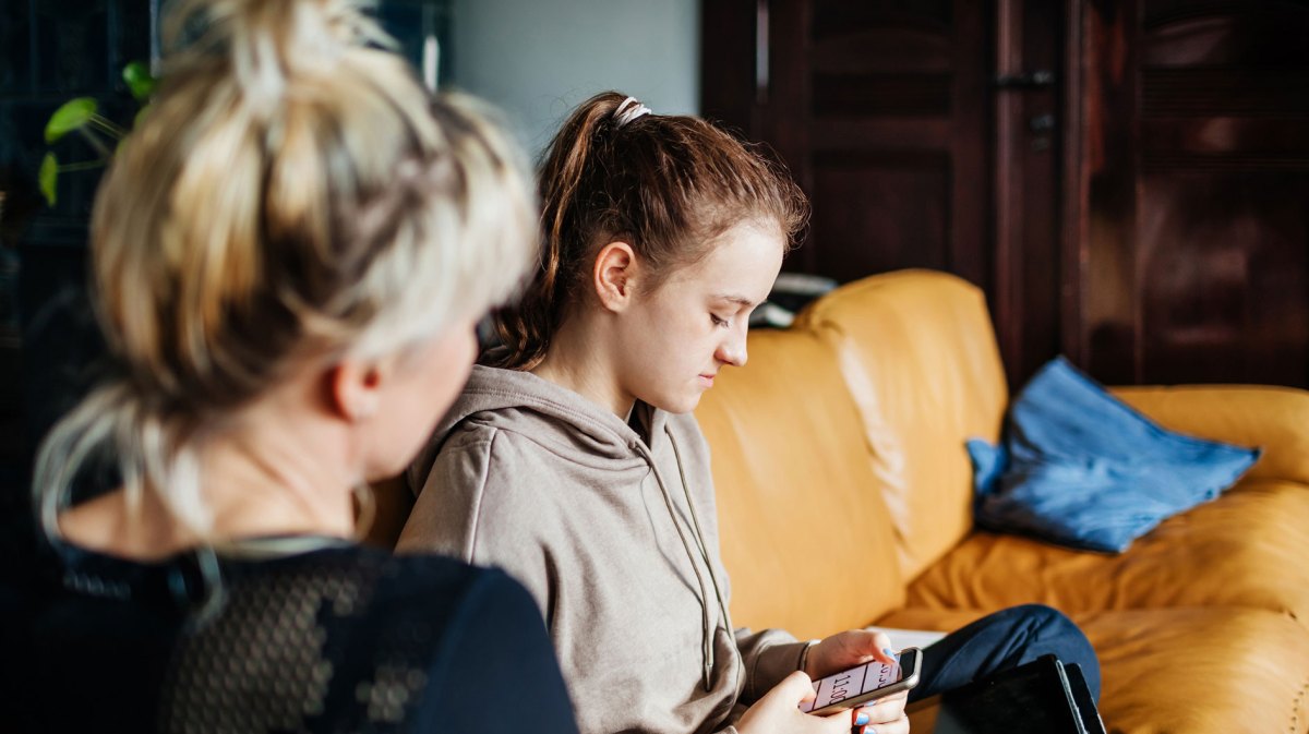 Mutter und Tochter auf der Couch. Die Tochter ist in ihr Handy vertieft, die Mutter versucht mimt ihr zu sprechen.