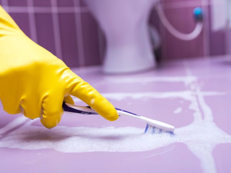 Hand mit gelbem Gummihandschuh putzt mit Zahnbürste Silikonfugen zwischen lila Fugen